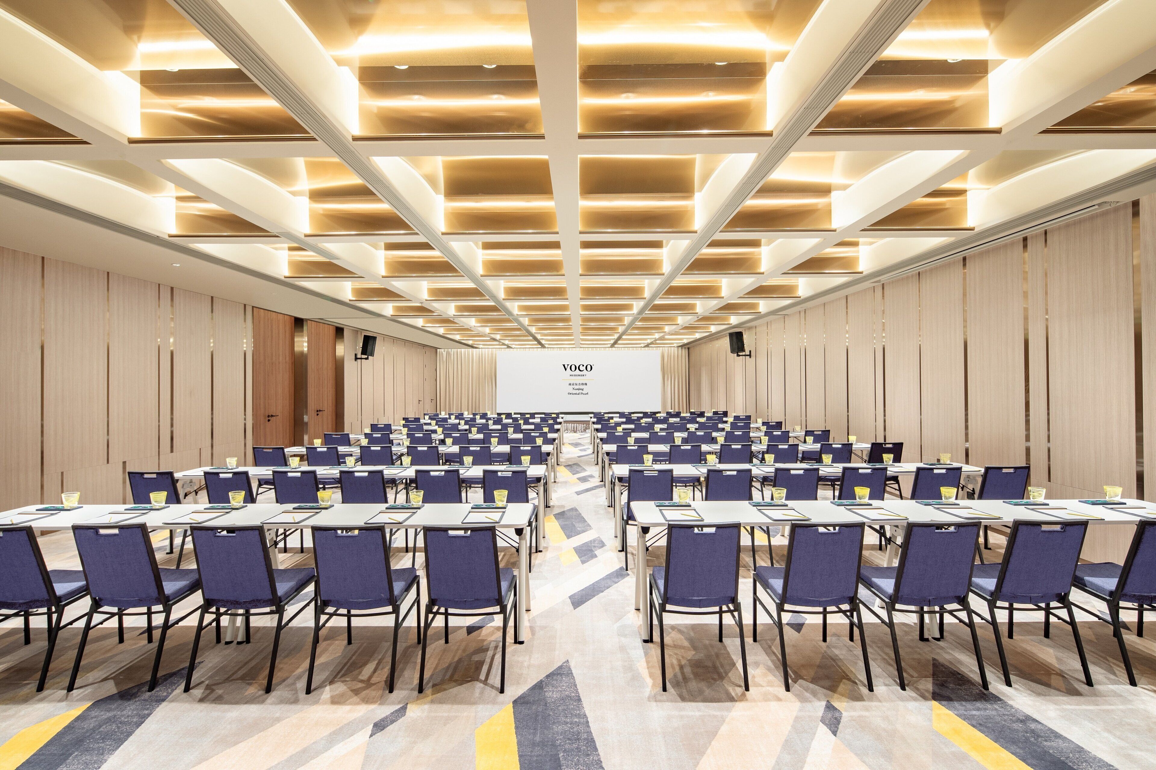 Un salón de conferencias con filas de mesas y sillas, un atril y una pantalla grande al frente.