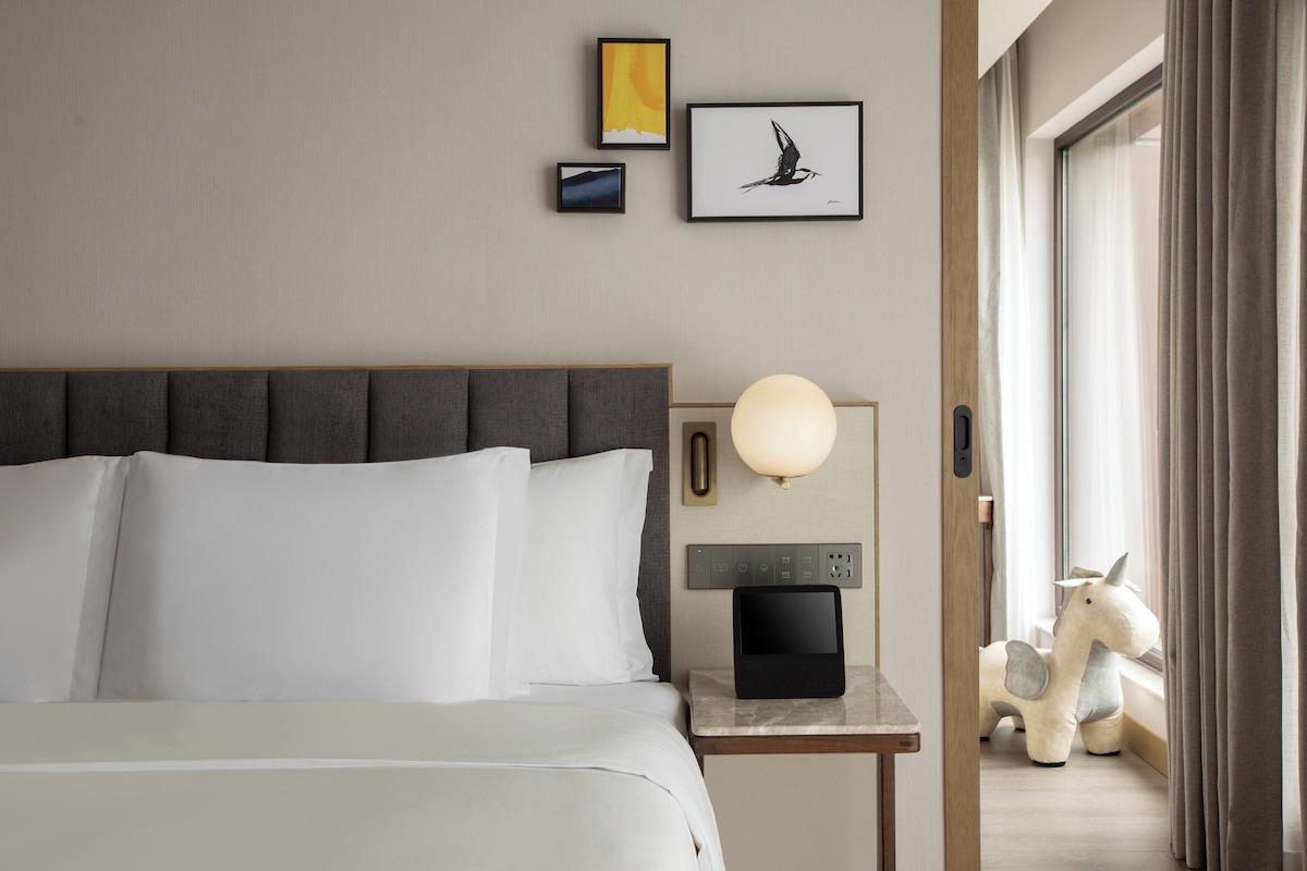 Ein Hotelzimmer mit einem Bett, einem Nachttisch mit Lampe, einer Bedienfeld und einem Kunstwerk an der Wand.