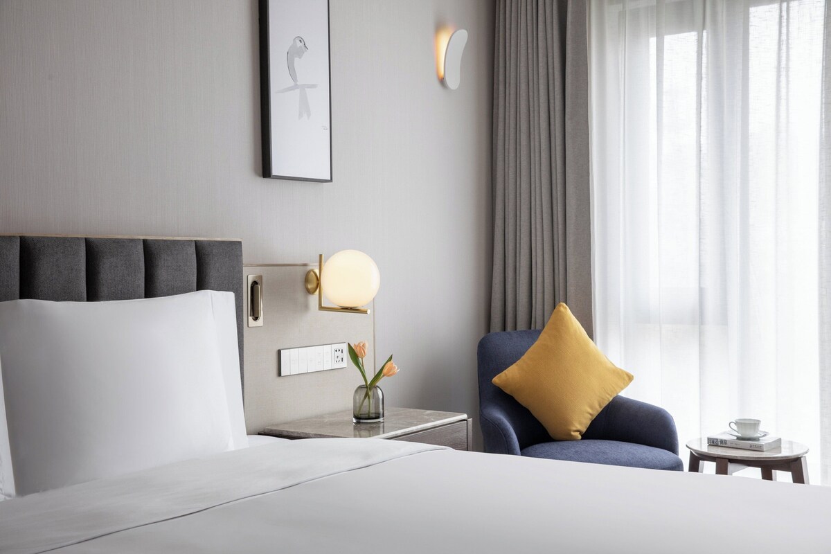 A hotel room with a bed, a chair, a nightstand, a lamp, a vase with flowers, and a window with curtains.