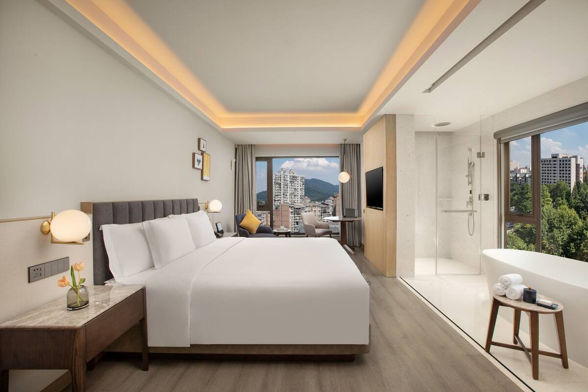 A modern hotel room with a large bed, a sitting area with a view, a separate bathroom, and a cityscape view.