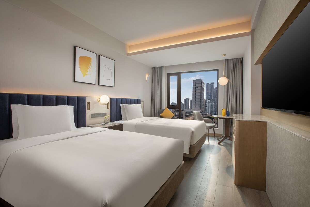 A hotel room with two beds, a desk, a TV, and a city view.