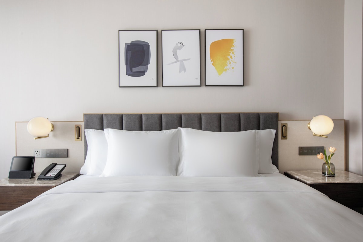 A hotel room with a large bed, two bedside tables with lamps, a phone, and three framed artworks on the wall.