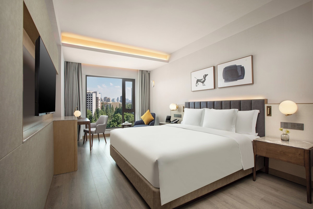Ein Hotelzimmer mit einem großen Bett, einem Schreibtisch, einem Sessel, einem Fernseher und Blick auf die Stadt.