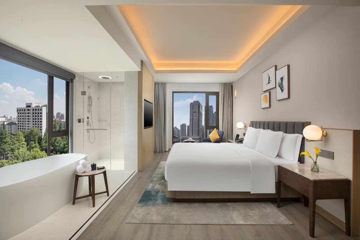 A modern hotel room with a large bed, a sitting area, a bathtub, and a view of the city skyline.