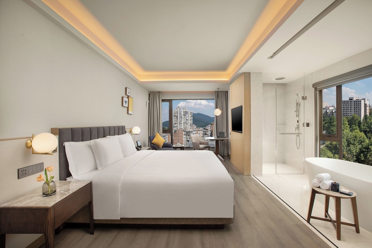 Ein Hotelzimmer mit Bett, Nachttisch, Lampe, Fernseher und Blick auf die Stadt.