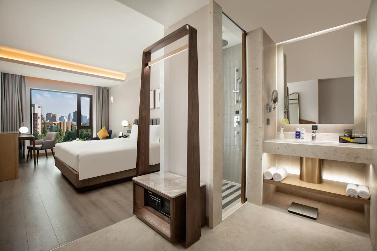 A modern hotel room with a large bed, a bathroom with a glass shower, and a city view.