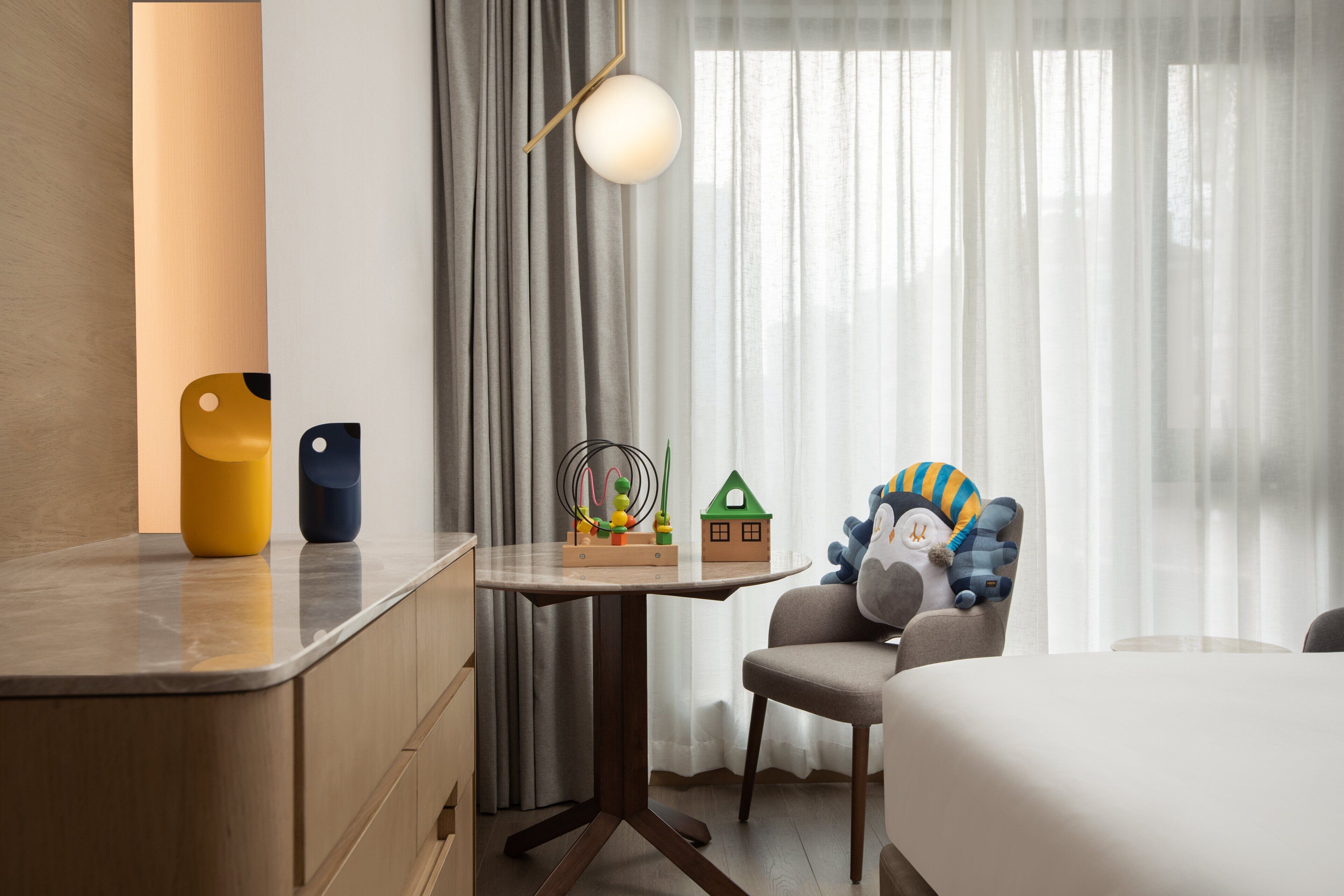 Una habitación de hotel con cama, escritorio con lámpara, silla con juguetes y ventana con cortinas.