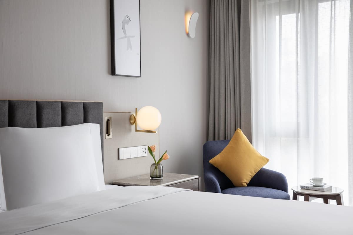 A hotel room with a bed, a nightstand with a lamp, a chair with a pillow, a vase with flowers, and a window with curtains.