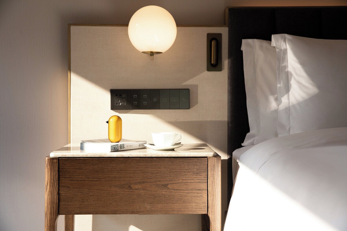 Ein Hotelzimmer mit einem hölzernen Nachttisch, einer Lampe, einer Tasse und einem Buch.