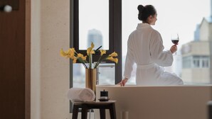 Una persona vestida con una bata blanca sentada junto a una ventana con vistas a edificios, sosteniendo una copa de vino tinto y un jarrón con flores amarillas sobre una mesita.