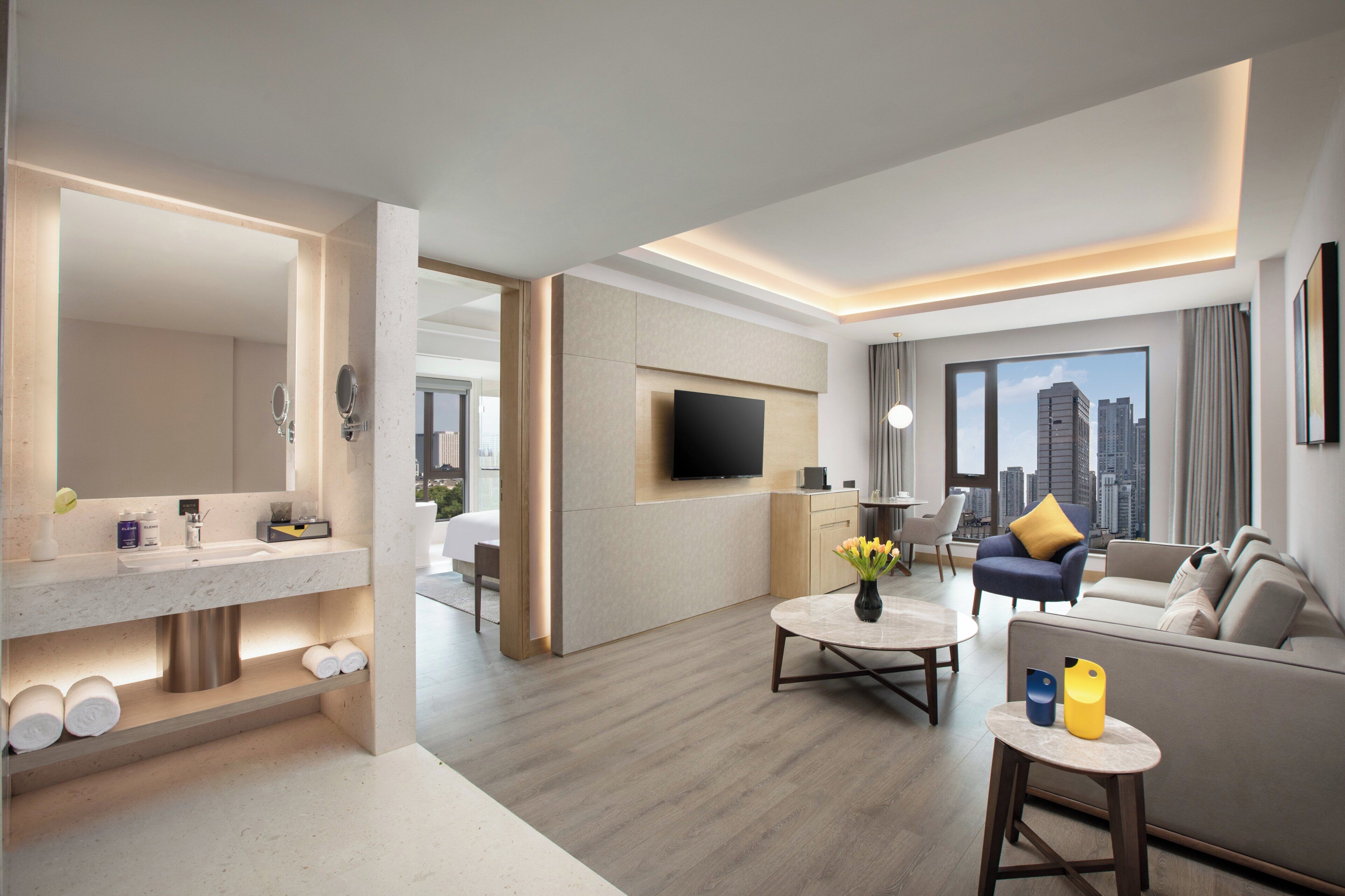 Habitación de hotel moderna con zona de estar, televisor de pantalla plana montado en la pared, sofá con cojín amarillo, mesa de centro con un jarrón de flores y un ventanal con vistas a la ciudad.