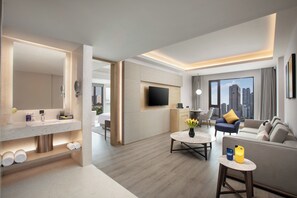A modern hotel room with a living area, a flat-screen TV mounted on the wall, a sofa with a yellow pillow, a coffee table with a vase of flowers, and a large window offering a city view. - voco Nanjing Oriental Pearl by IHG (Nanjing)