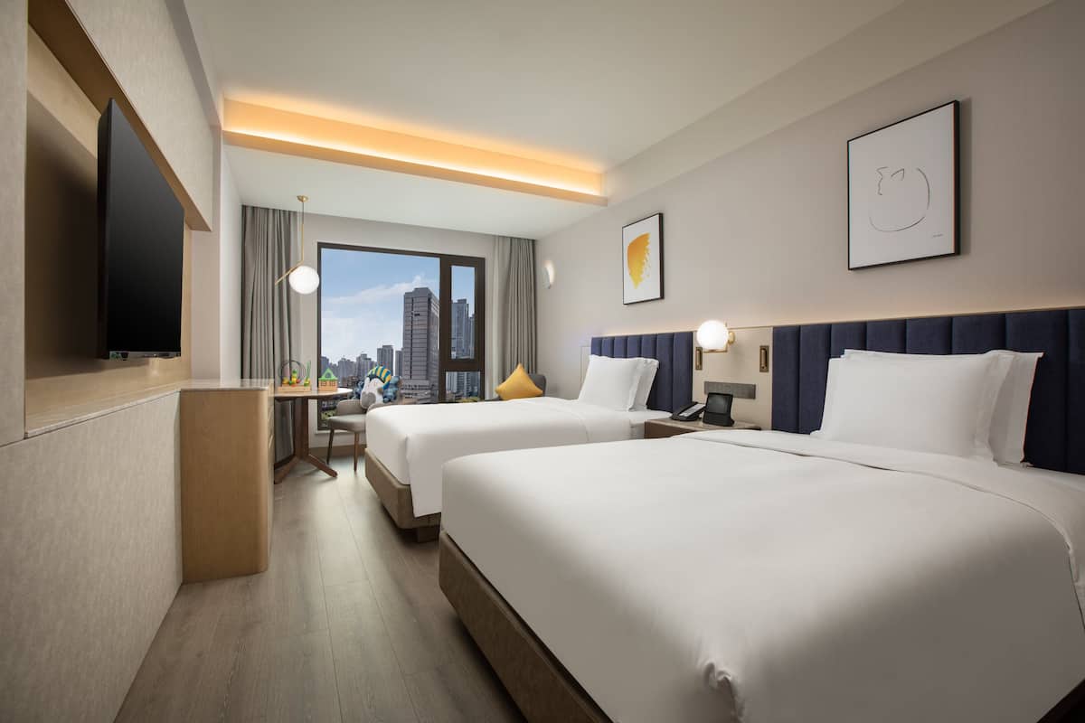 A hotel room with two beds, a desk, a flat-screen TV, and a city view.