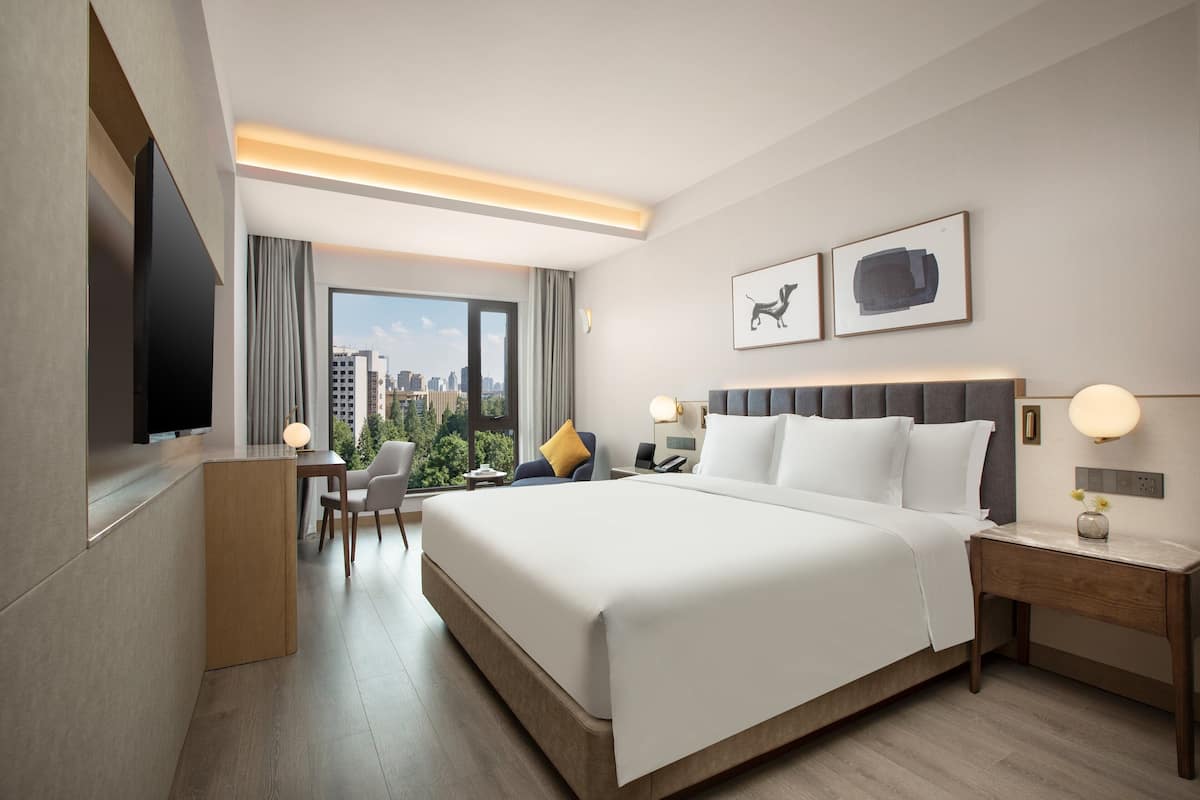 A modern hotel room with a large bed, a desk, a chair, a TV, and a city view.