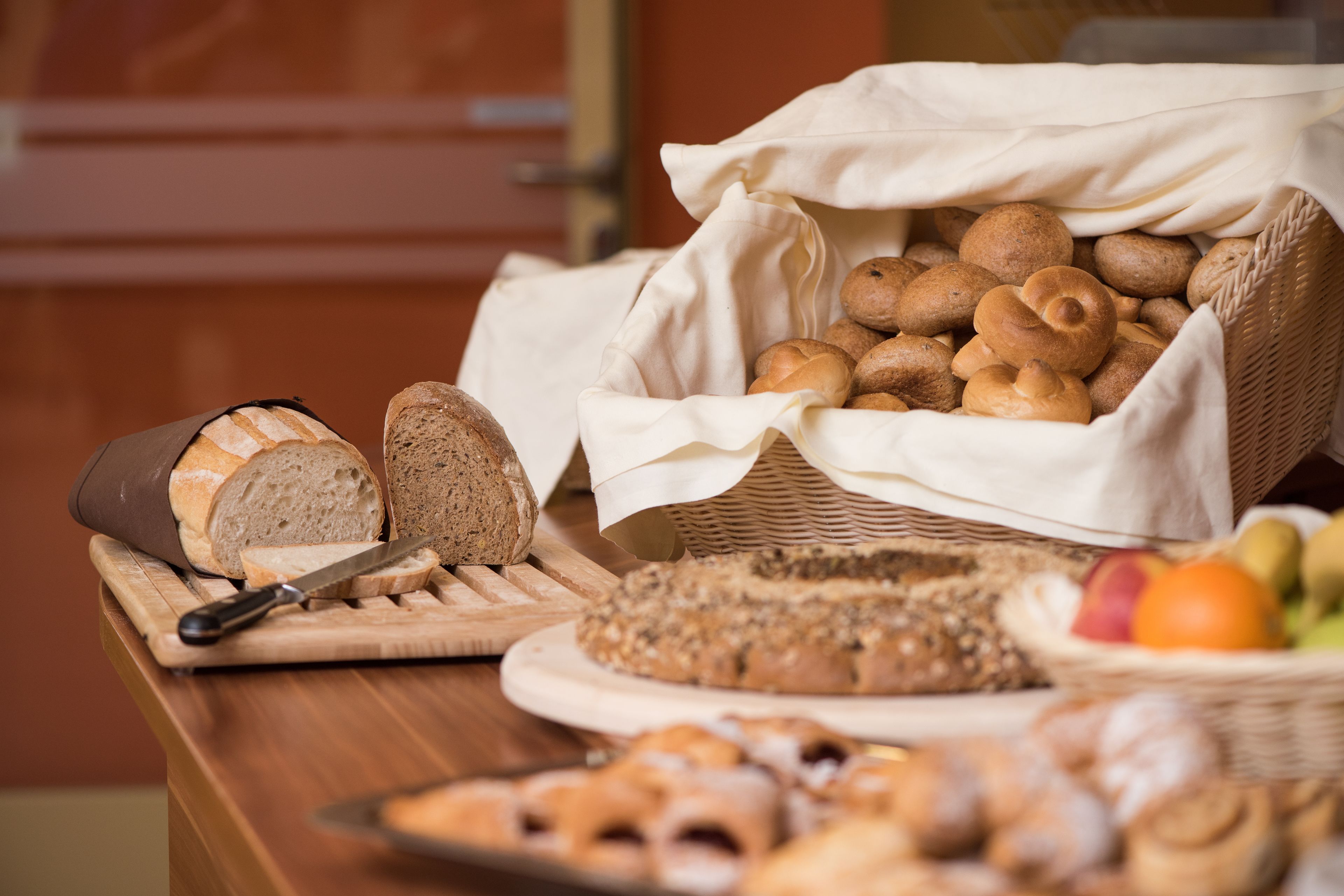 Variedad de panes y pasteles sobre una mesa de madera, incluyendo una hogaza, rebanadas de pan y una cesta de bollos redondos.