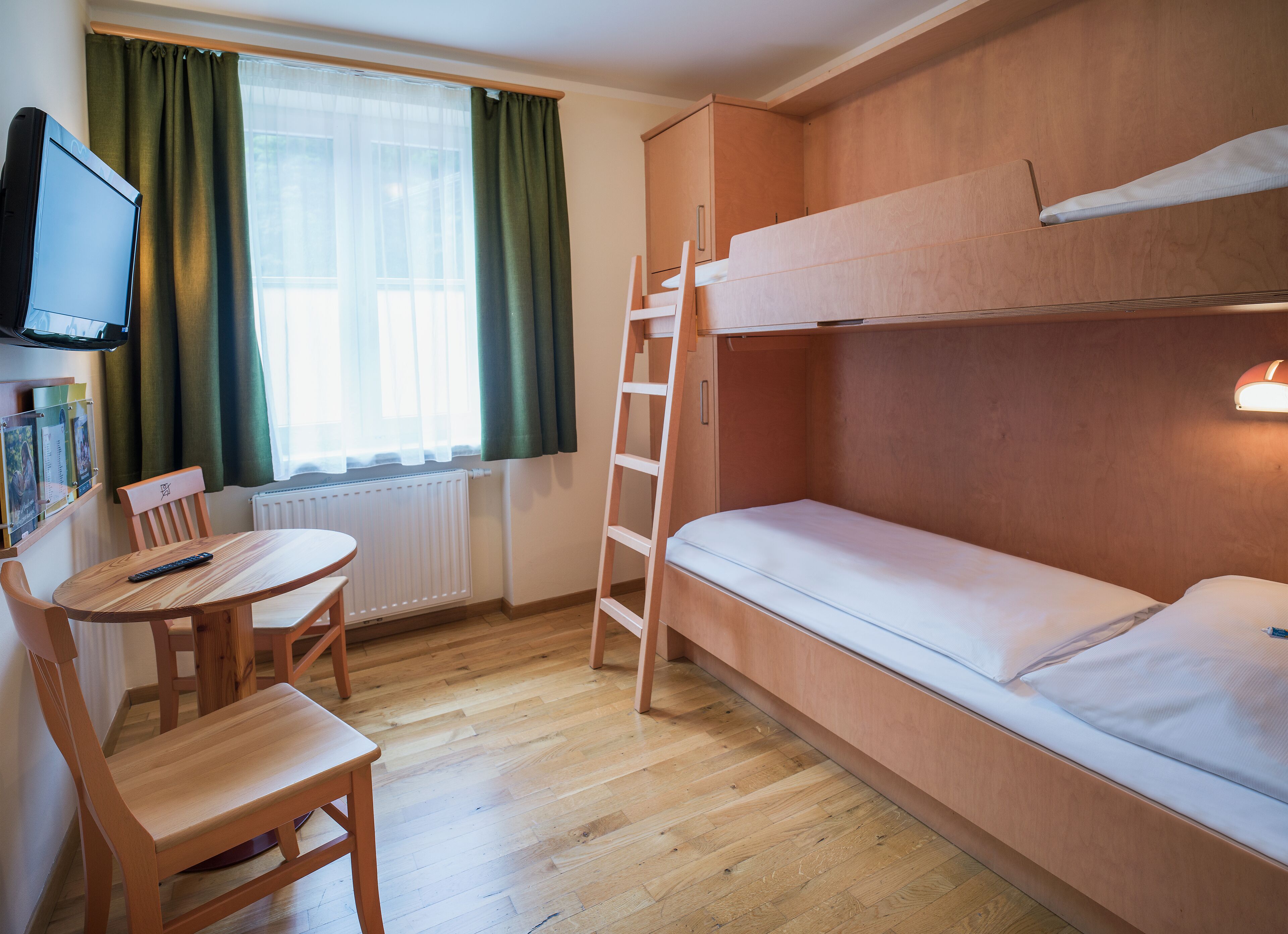 Una stanza compatta con un letto a castello, un tavolino con una sedia, una TV fissata al muro e una finestra con tende.