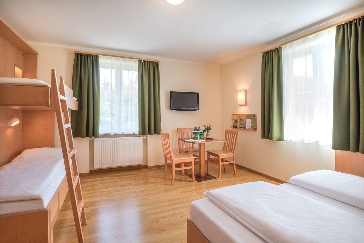 A hotel room with a bunk bed, a desk with chairs, a television, and a window with curtains.