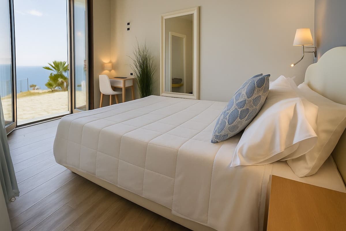 A bedroom with a large bed, a bedside table, a chair, a small table, a lamp, and a view of the beach.