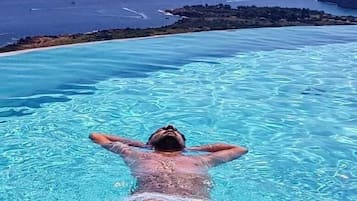 A person swimming in a clear blue infinity pool with a view of the ocean and distant mountains.