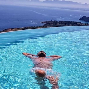 A person swimming in a clear blue infinity pool with a view of the ocean and distant mountains. - L'Antica Cascina del Golfo (Castellammare del Golfo)