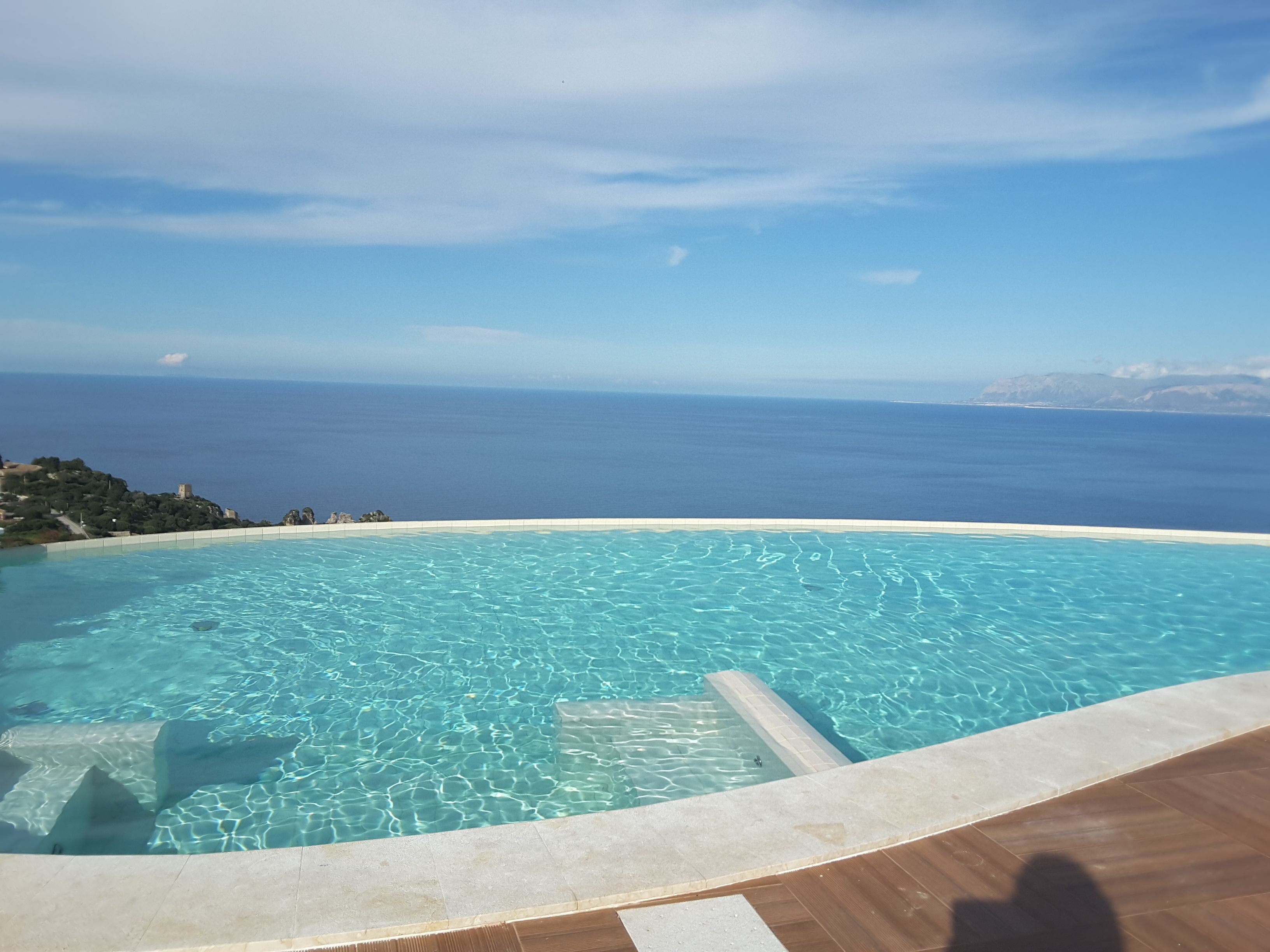 Una piscina infinita con agua azul cristalina, una terraza de madera y vistas al océano y montañas distantes.