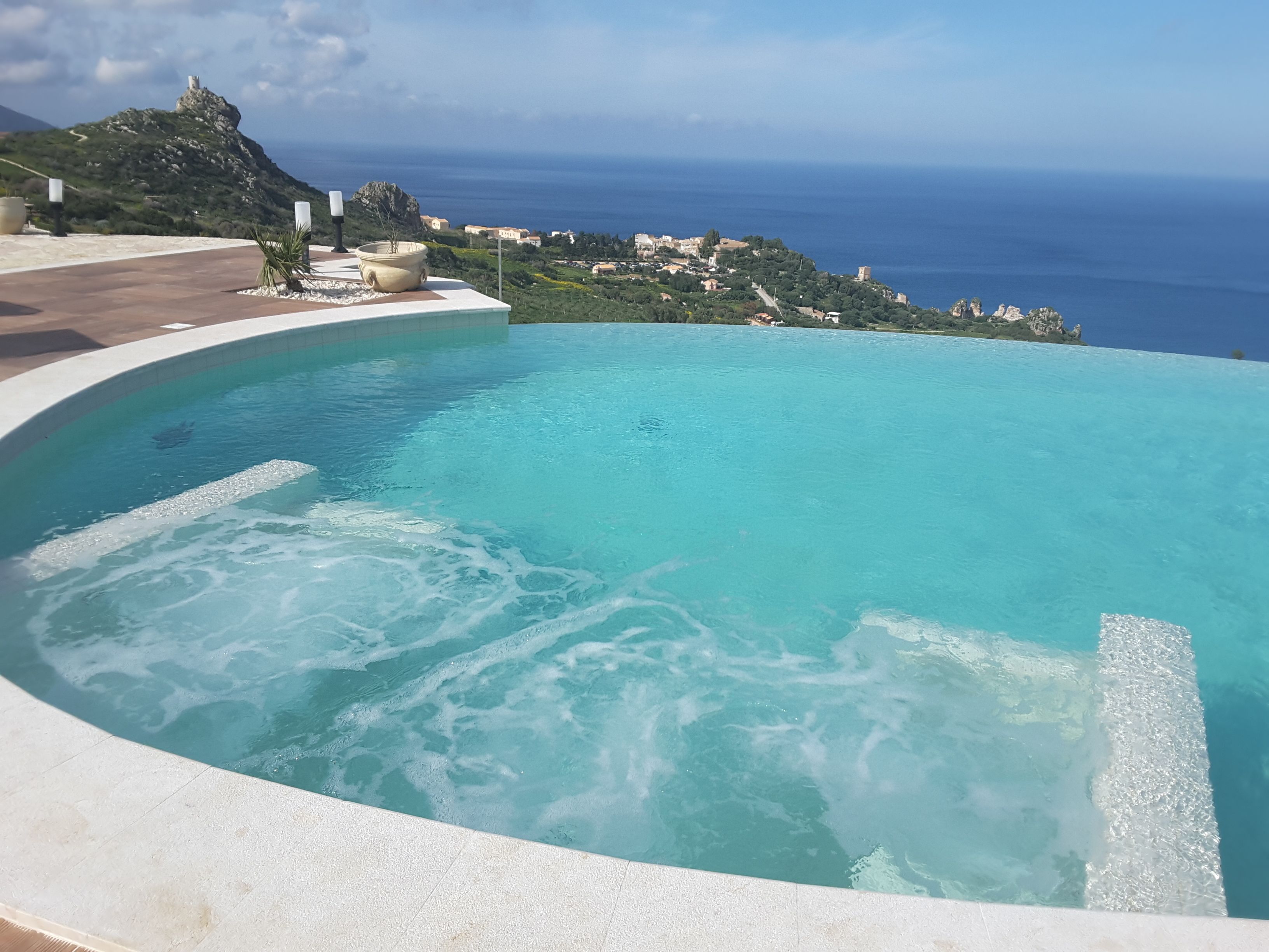 A swimming pool with a curved edge, surrounded by a paved area with a view of the sea and a distant town.
