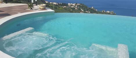 A swimming pool with a curved edge, surrounded by a paved area with a view of the sea and a distant town.