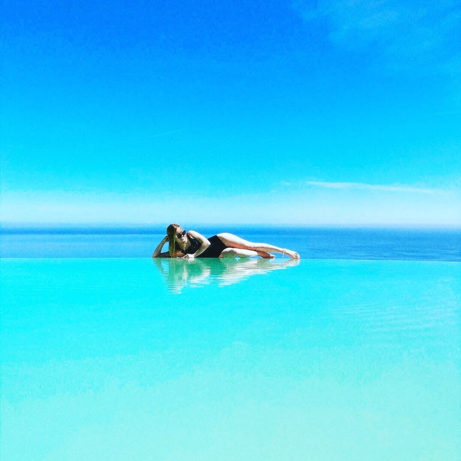 A person lying on a clear blue water surface, likely a pool, with a vast ocean and clear sky in the background.
