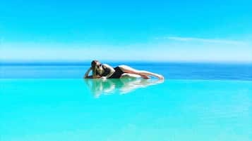 A person lying on a clear blue water surface, likely a pool, with a vast ocean and clear sky in the background.