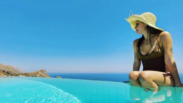 Una mujer sentada al borde de una piscina infinita azul y transparente, con vista al océano y colinas distantes.