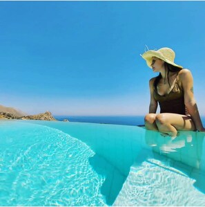 A woman sitting on the edge of a clear blue infinity pool with a view of the ocean and distant hills. - L'Antica Cascina del Golfo (Castellammare del Golfo)