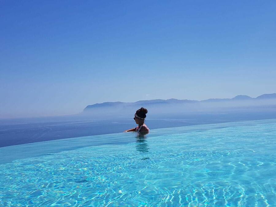 Uma pessoa à beira de uma piscina azul cristalina com vista para montanhas ao longe.