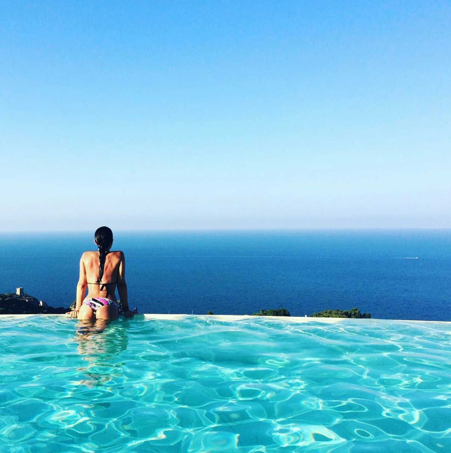 A person in a bikini sitting at the edge of a clear blue pool with a view of the ocean.