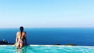 A person in a bikini sitting at the edge of a clear blue pool with a view of the ocean.