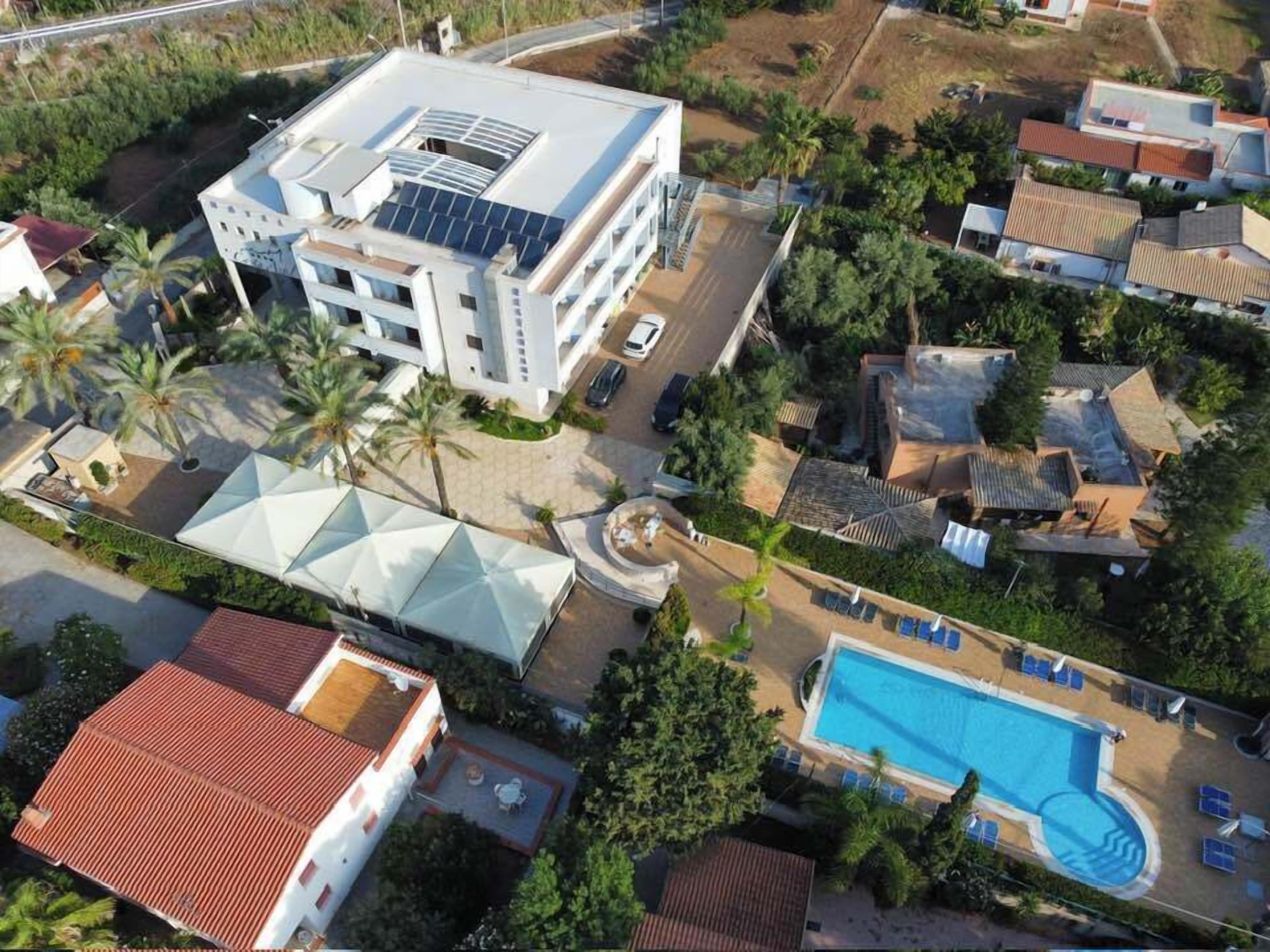 An aerial view of a residential area with a modern building featuring a rooftop solar panel array, a swimming pool with sun loungers, and surrounding houses with red-tiled roofs.
