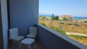 A balcony with two white chairs, a view of a coastal area with houses and mountains in the distance.