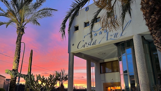 Entrada de un hotel con una palmera y cactus en primer plano, y un atardecer al fondo.