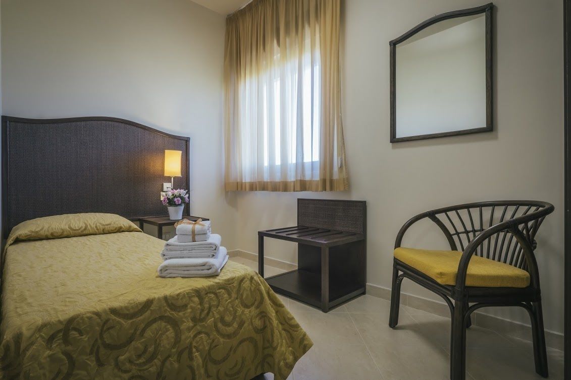 A hotel room with a bed, a chair, a small table with a lamp, and a window with curtains.