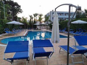 A poolside area with blue lounge chairs and umbrellas, surrounded by palm trees and a building in the background.