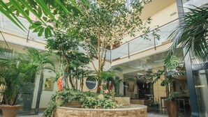 Un atrio espacioso con varios niveles, que cuenta con un jardín central con diversas plantas y árboles, rodeado de barandillas de vidrio y arquitectura moderna.