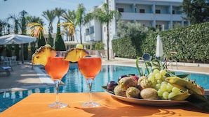 Escena junto a la piscina con dos cócteles, una bandeja de frutas y vistas a un edificio moderno.
