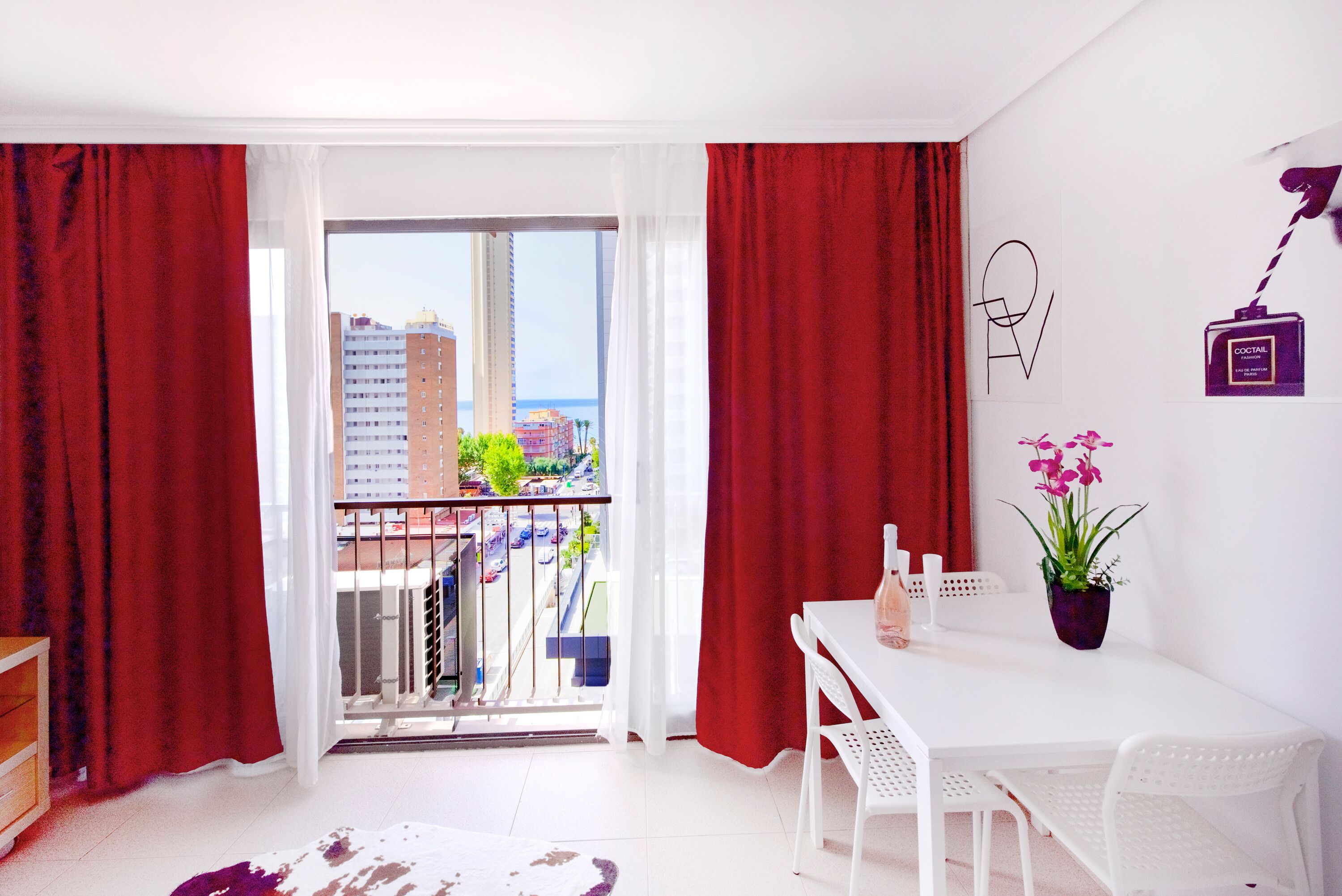 Una habitación con balcón, muebles blancos, una planta en maceta y un objeto montado en la pared.