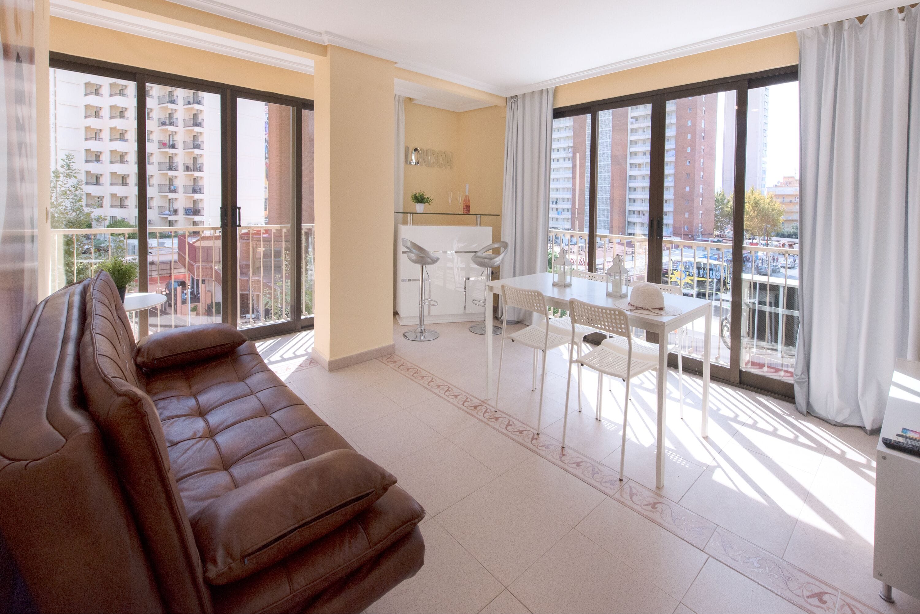A modern living room with a brown sofa, a dining area with a white table and chairs, and large windows offering a view of buildings and a street.