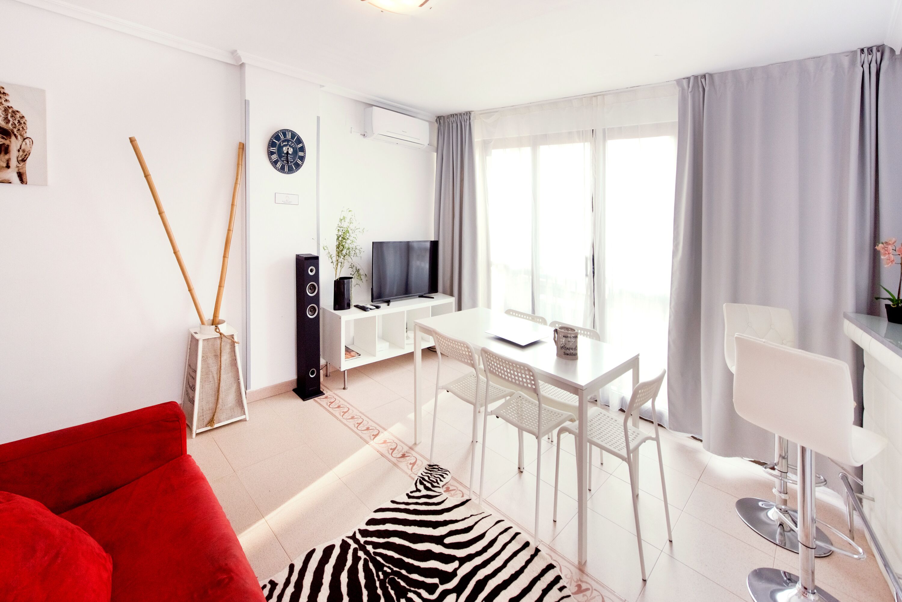 A modern living room with a red sofa, a zebra-patterned rug, a dining area with white chairs, and a TV on a stand.