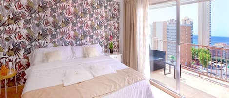 A hotel room with a large bed, a balcony with a view of the city, and a tropical wallpaper design.