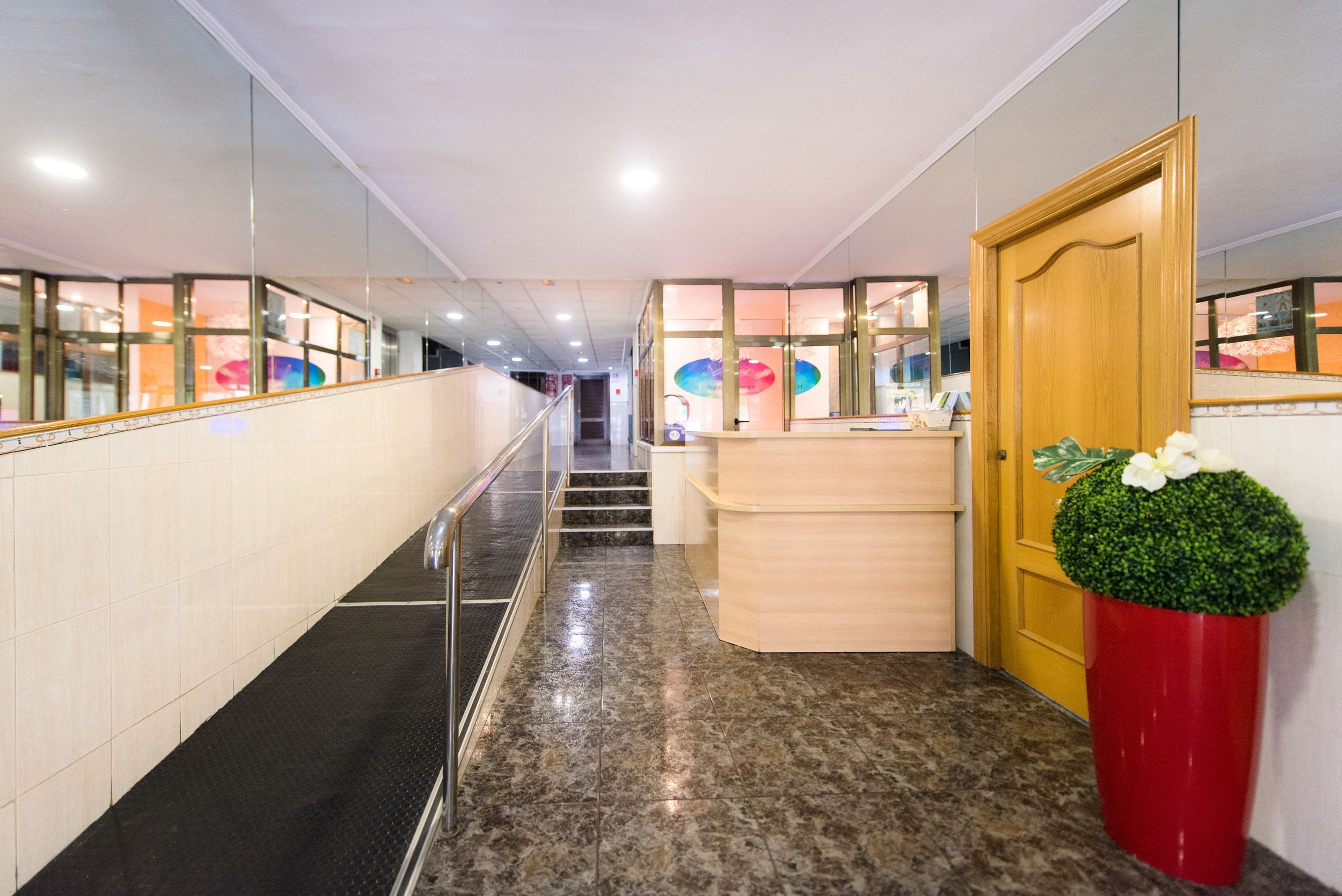 A hotel reception area with a marble floor, a wooden reception desk, a red flower pot, and a wooden door.