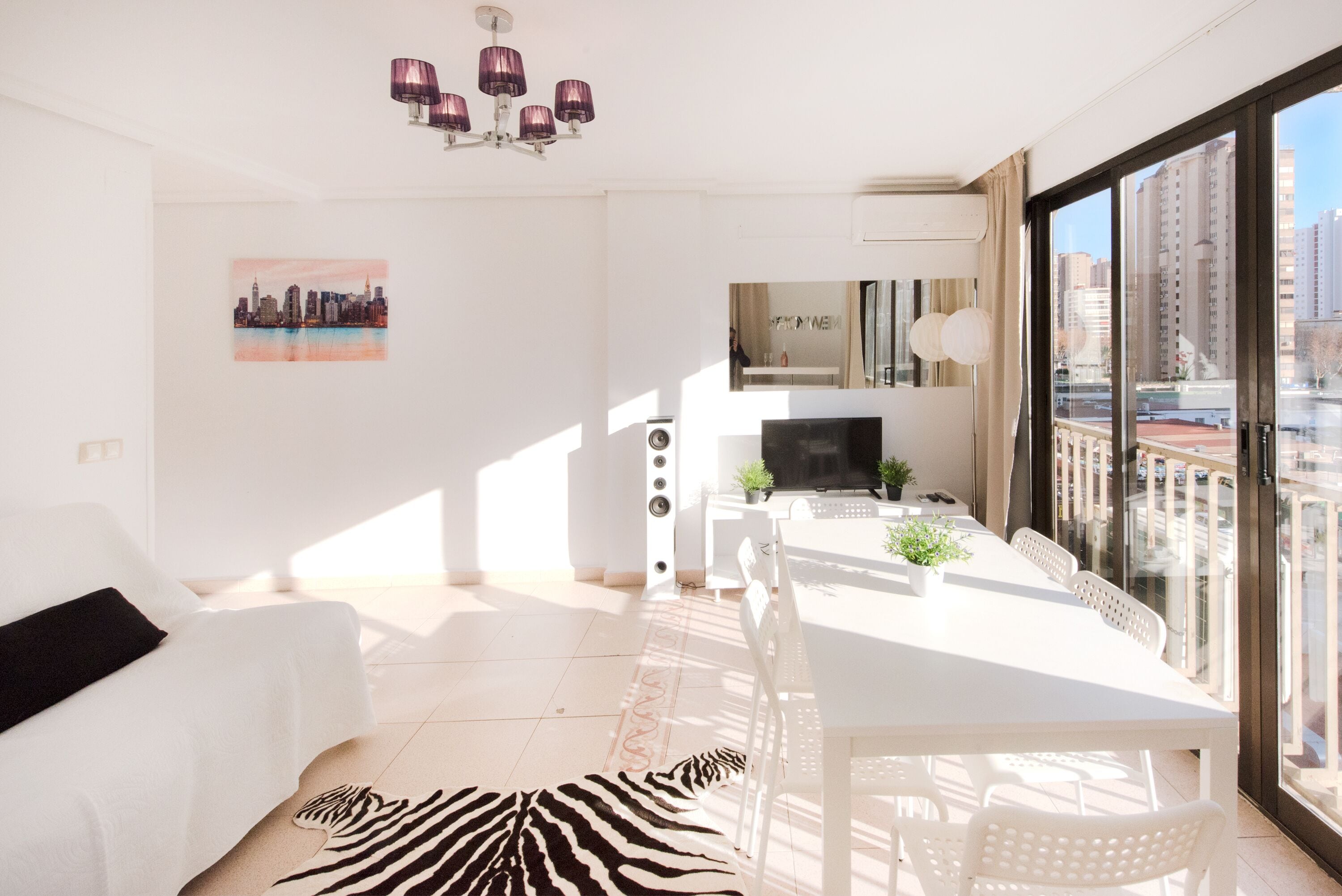 Un salon moderne avec un canapé blanc, un tapis à motifs zébrés, une table à manger avec des chaises, un téléviseur et une vue sur le paysage urbain à travers de grandes fenêtres.