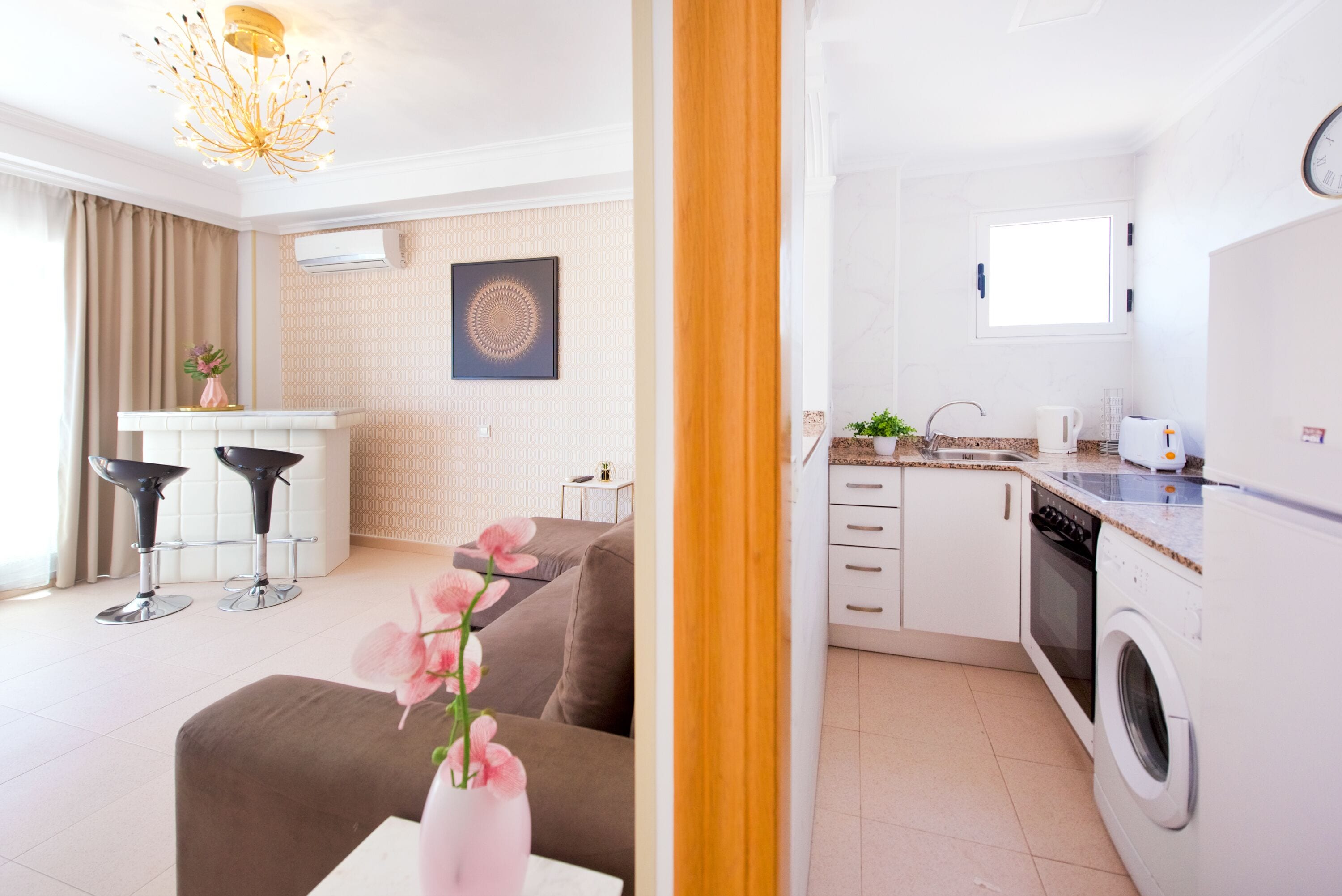 Una cocina moderna con lavadora, un comedor con dos taburetes tipo barra, y una sala de estar con sofá y un jarrón de flores.