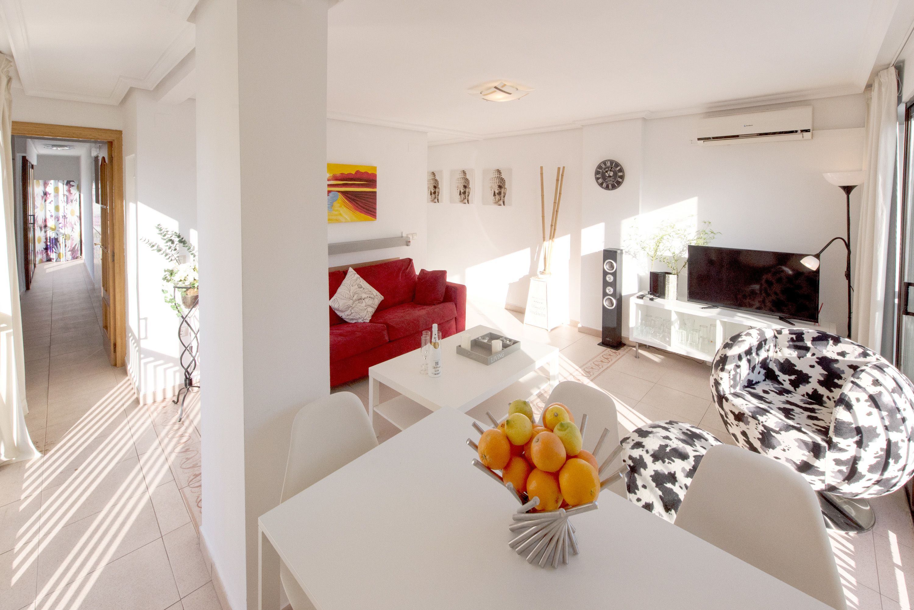 A modern living room with a red sofa, white coffee table, and a TV. There is a dining area with a white table and chairs, and a fruit bowl on the table.
