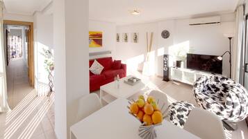 A modern living room with a red sofa, white coffee table, and a TV. There is a dining area with a white table and chairs, and a fruit bowl on the table.